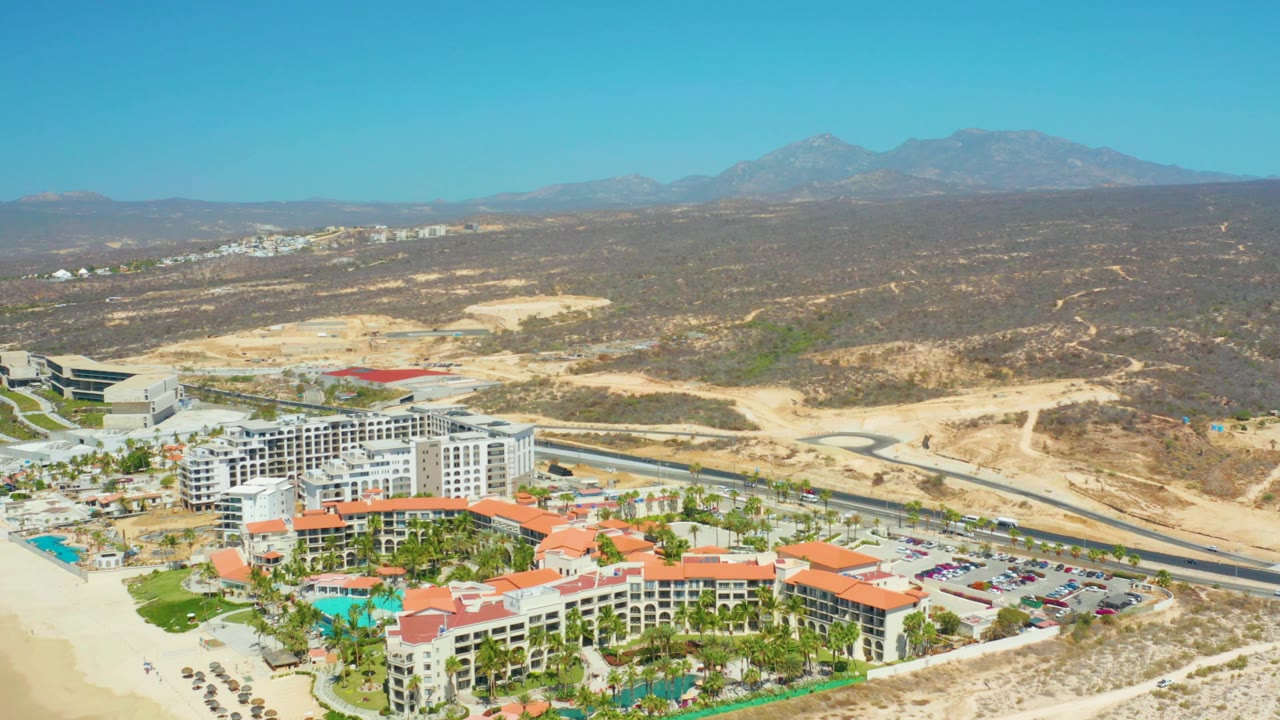 Aerial frame of the Dreams Los Cabos resort complex with red-tiled roofs and pool areas in the lower frame, parking and road in the middle distance, brown hills, mountains, and no visible people.