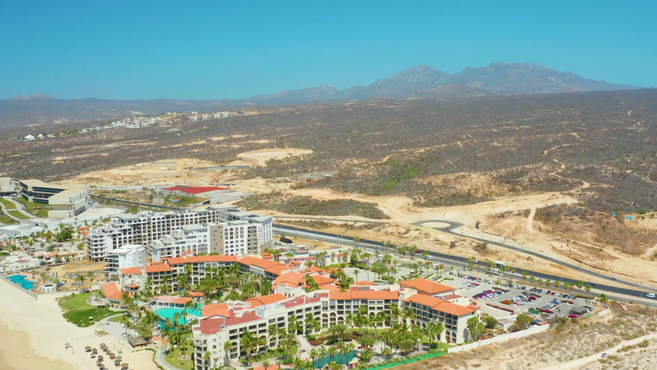 At 0:03, the Dreams Los Cabos resort remains in the lower foreground with red roofs, pools, beach edge, adjacent road, dry hillside terrain, distant mountains, and no visible people.