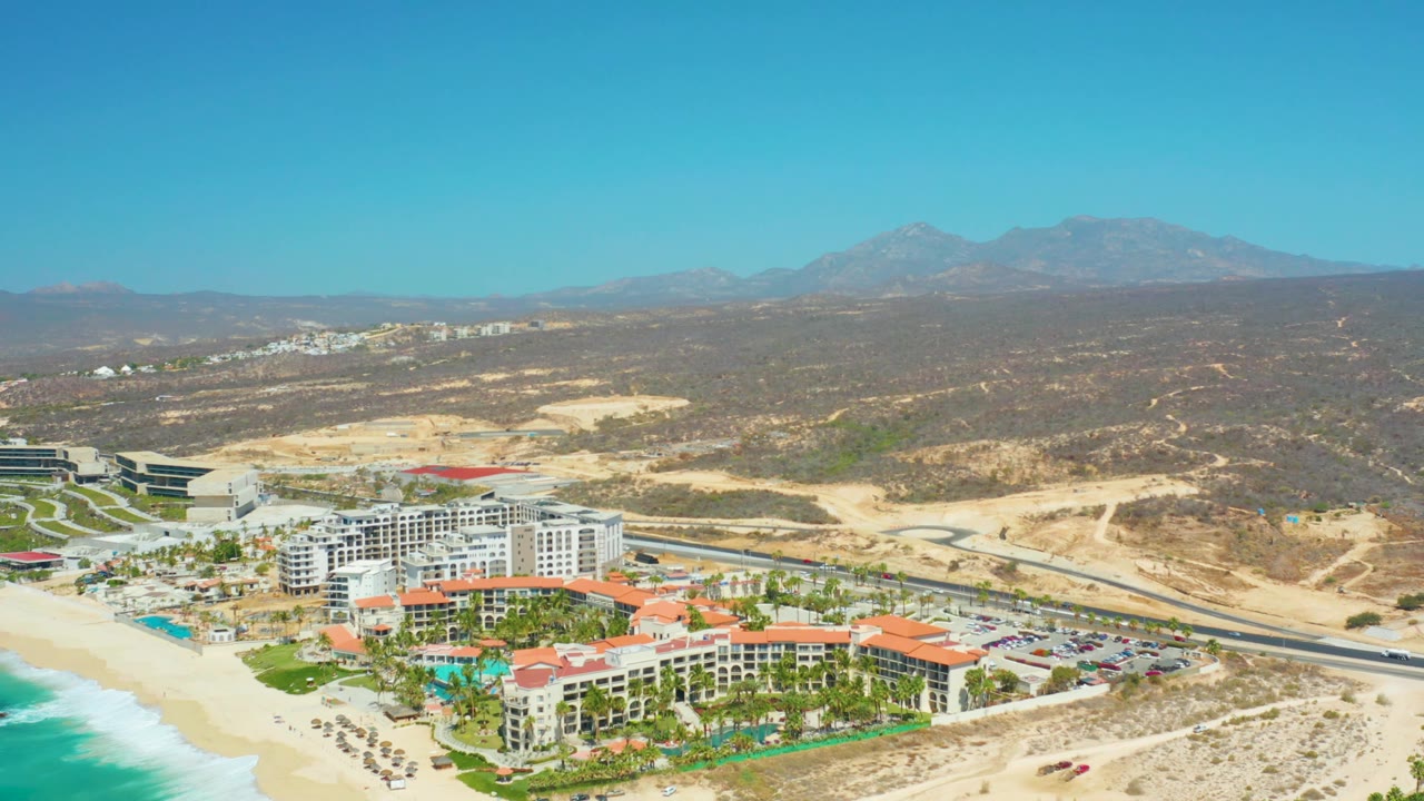 Aerial frame at Dreams Los Cabos with the resort slightly more centered, ocean surf at the lower left, road across the midground, desert hills behind, distant mountains, and no visible people.