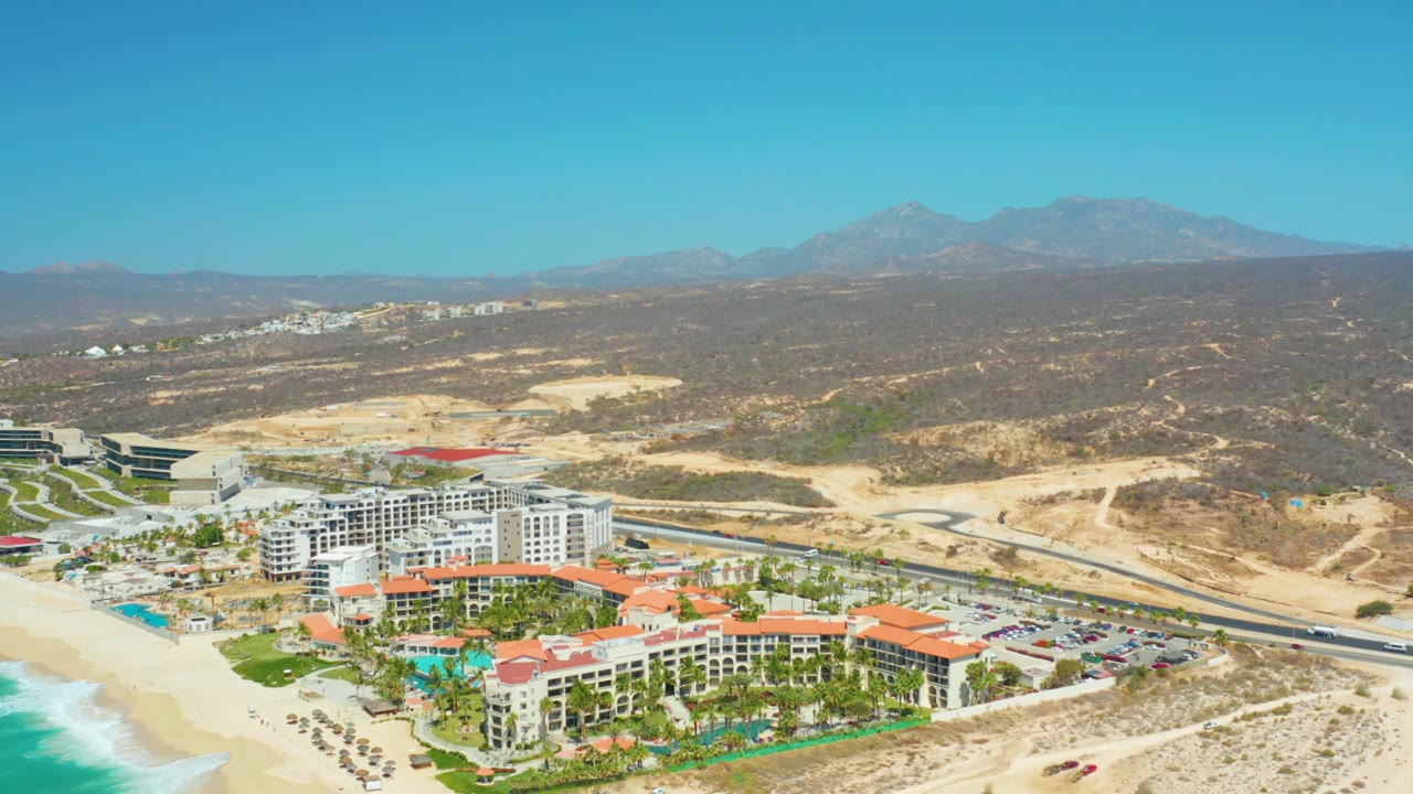 Mid-clip aerial frame over Dreams Los Cabos showing beach and ocean at lower left, resort pools and buildings below, the road crossing the scene, dry hills behind it, mountains, and no visible people.