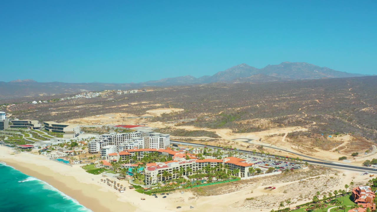 Dreams Los Cabos aerial view with more of the beachfront resort centered, small beach umbrellas near the sand, the road behind the buildings, and mountains across the horizon.