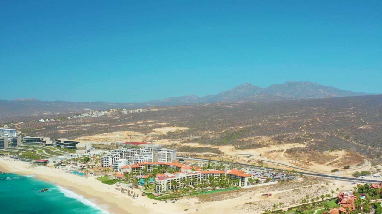 Aerial view at 0:01 over Dreams Los Cabos, with the beach and ocean at lower left, clustered resort buildings near the shore, the highway crossing the middle distance, and no visible people.