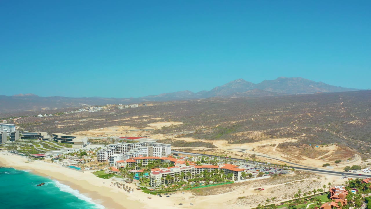 A 0:02 aerial frame of Dreams Los Cabos where the sandy shoreline, white resort structures, highway curve, scrubby brown terrain, and distant mountain range remain clearly visible with no visible people.