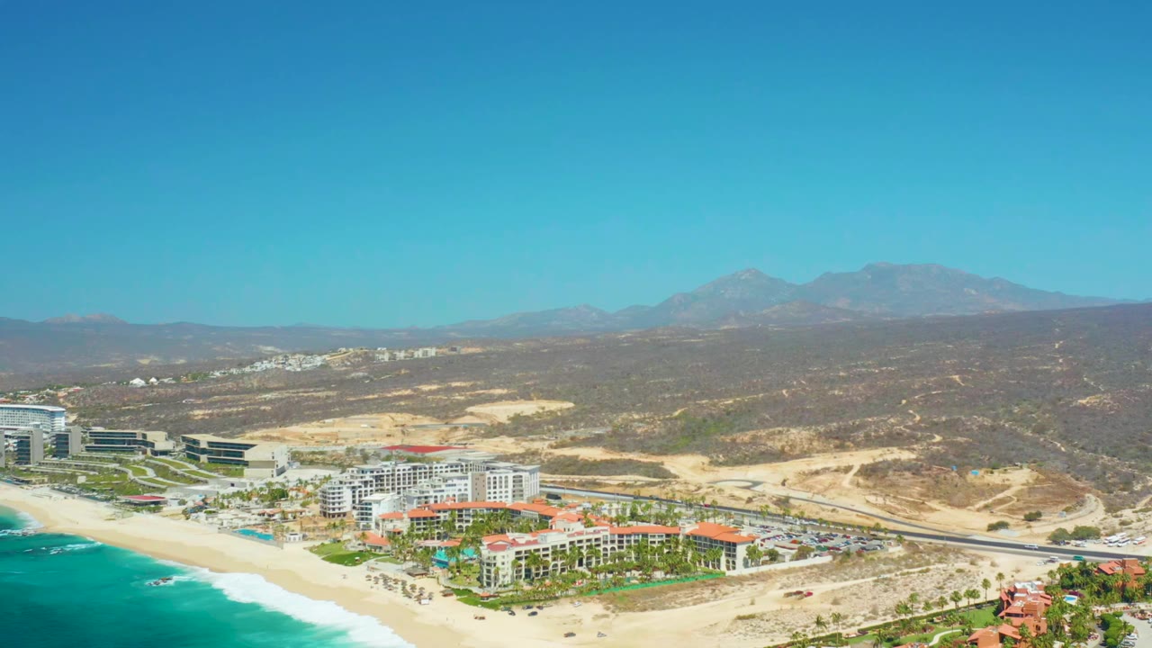 At 0:01, the Dreams Los Cabos resort remains centered near the beach, with white surf along the lower left edge, tan hills behind the buildings, mountains on the horizon, and no visible people.