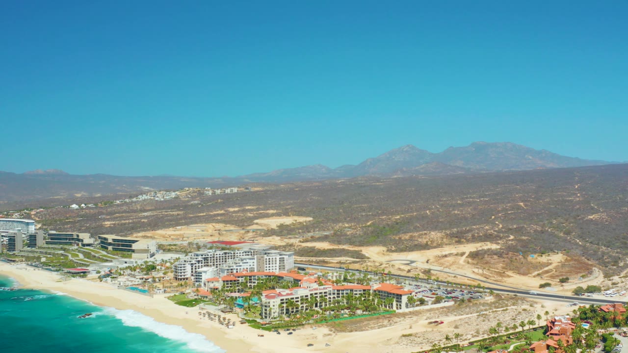At 0:03, Dreams Los Cabos appears from the same elevated coastal angle, with the beach and turquoise sea at lower left, resort blocks and grounds in the midground, tan hills beyond, and no visible people.