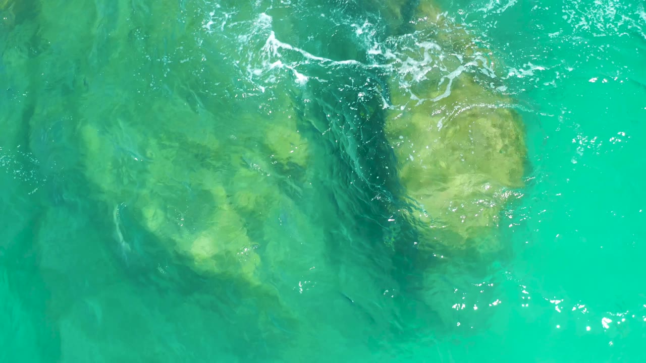 At 0:03, pale rocks are visible beneath turquoise water while a white foam line crosses near the upper left, with no shoreline, boats, or visible people.
