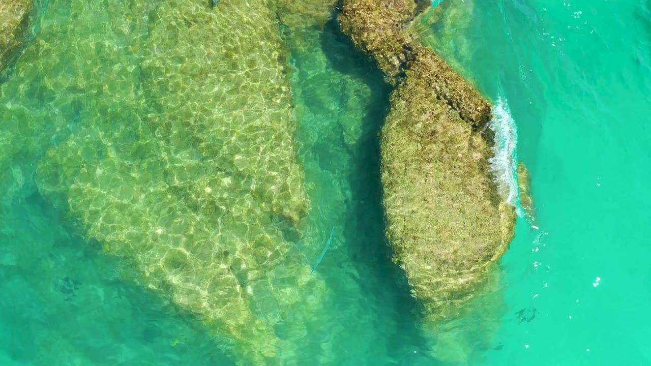 Overhead frame from Dreams Los Cabos showing clear turquoise water, submerged tan rock shelves, a darker narrow channel, and no visible people.