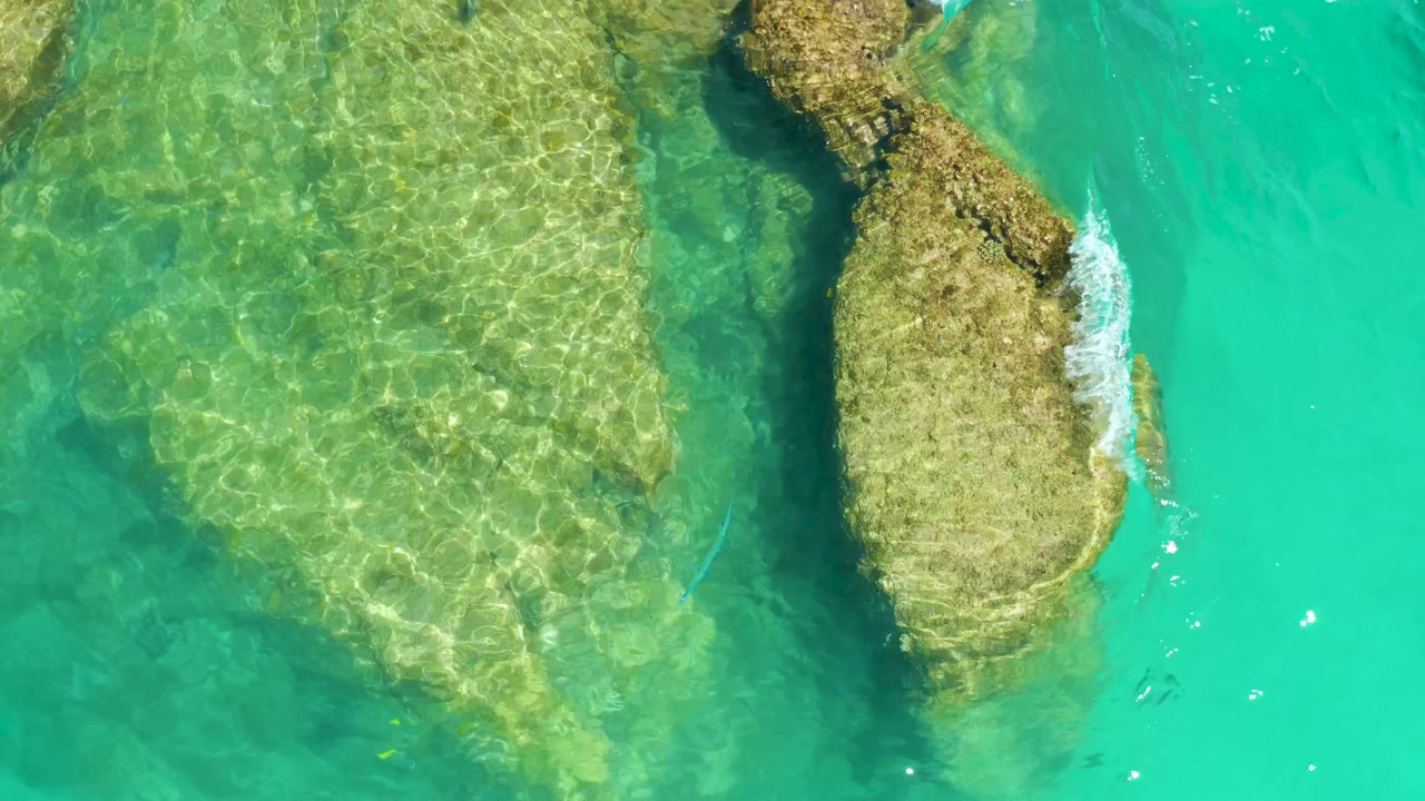 At 0:01, the overhead view holds on the shallow rocky seabed at Dreams Los Cabos, with bright turquoise water and no visible people.