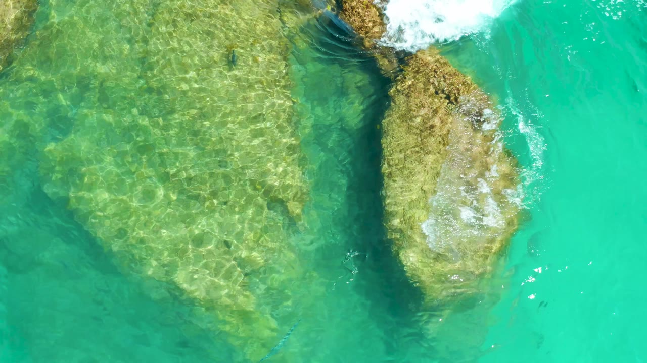 At 0:02, white wave foam enters from the upper right over the exposed rock edge in the Dreams Los Cabos shallows, with no visible people.