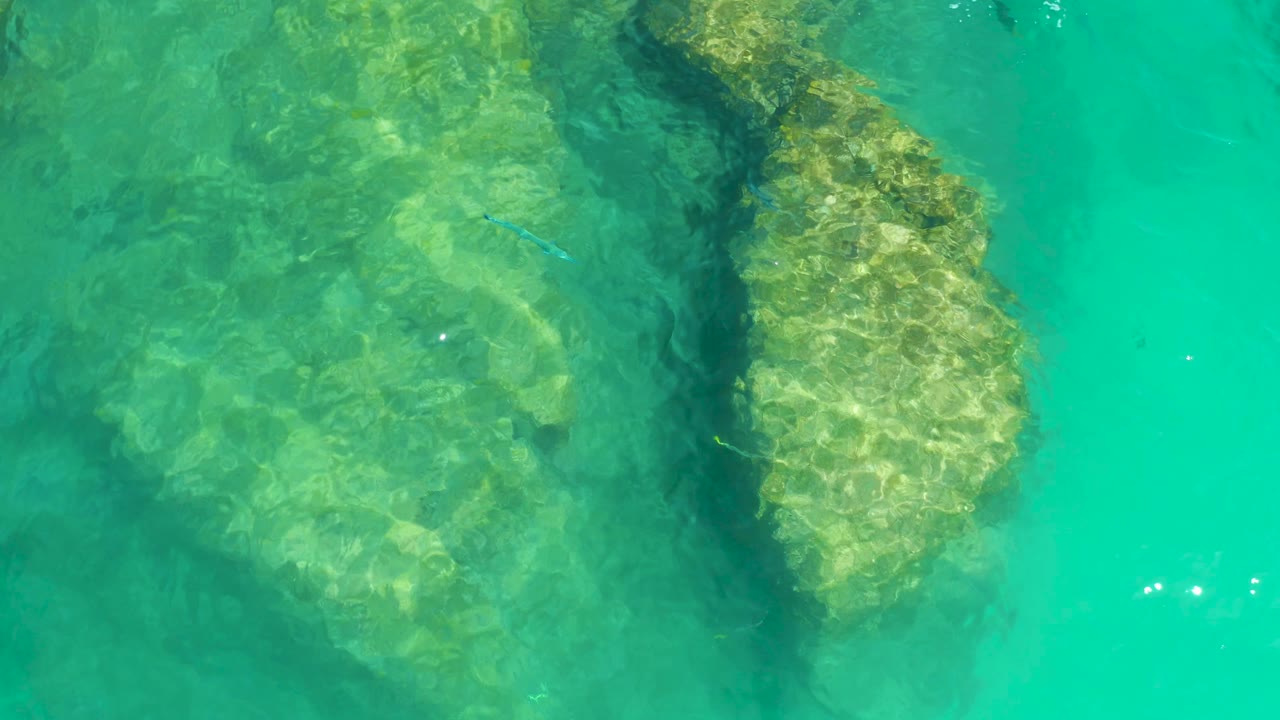 Clear water at Dreams Los Cabos reveals pale submerged rock shapes, a darker crevice, small fish over the seabed, bright surface glints, and no visible people.