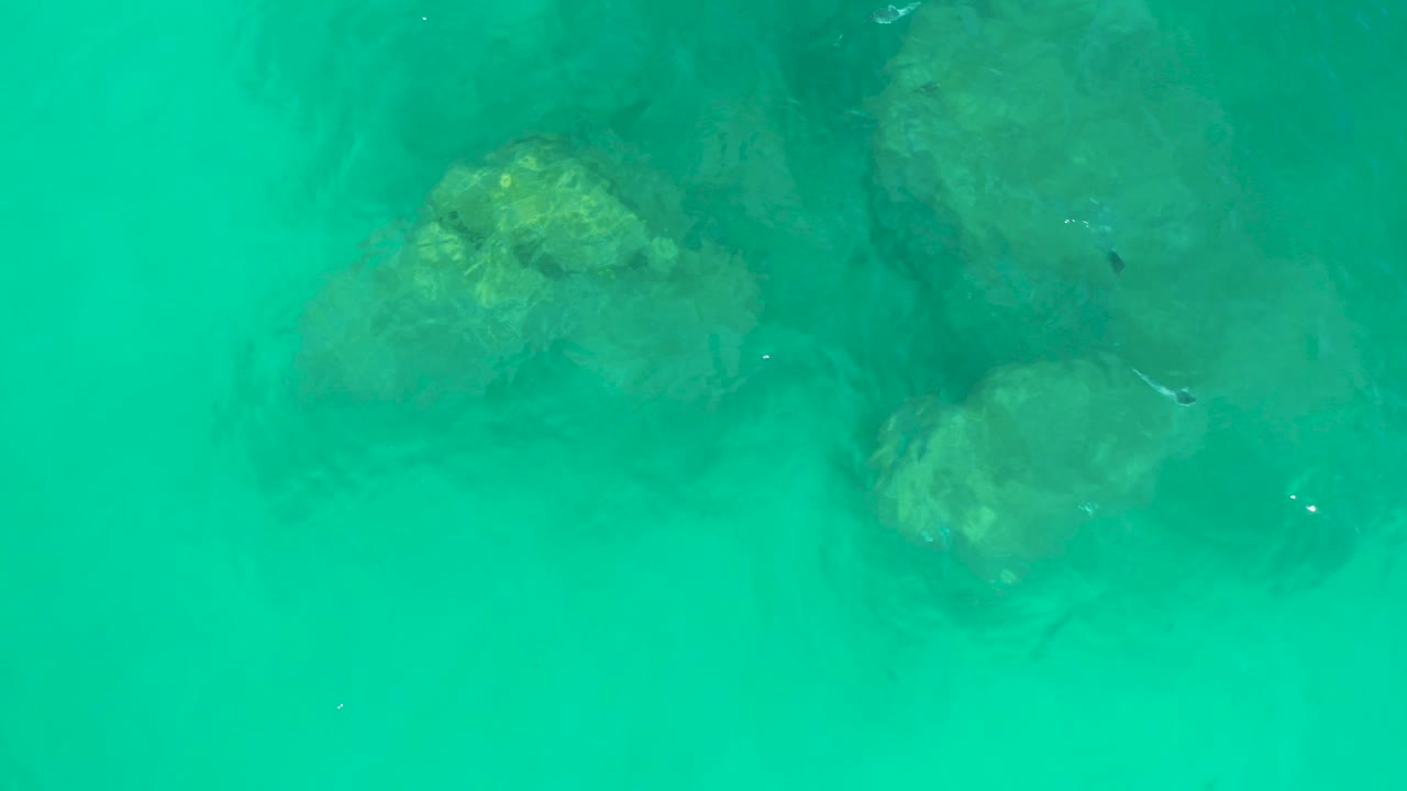 Overhead view of turquoise water at Dreams Los Cabos with submerged rocks near the upper frame, sandy shallows below, faint fish shapes, and no visible people.