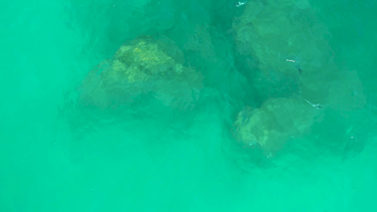 A slightly shifted overhead frame shows green-blue water over clustered submerged rocks at Dreams Los Cabos, with lighter sand around the rocks and no visible people.