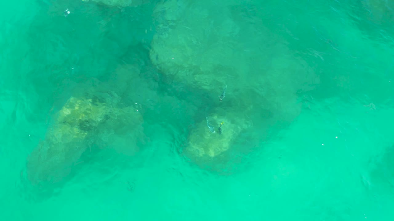 At 0:03, the downward view shows clear shallow ocean water with several pale rocks beneath shifting green surface reflections, with no visible people.