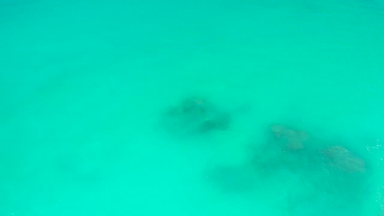 Clear turquoise water at Dreams Los Cabos with a darker rock cluster high in frame and a larger seabed formation to the right, no visible people.