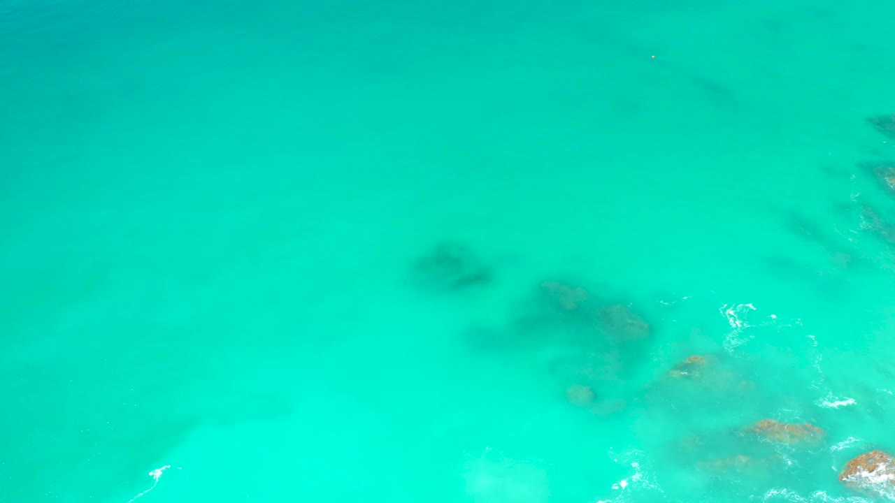 Clear blue-green water at Dreams Los Cabos covers dark and brown seabed rocks, with a small bright foam streak near the lower left and no visible people.