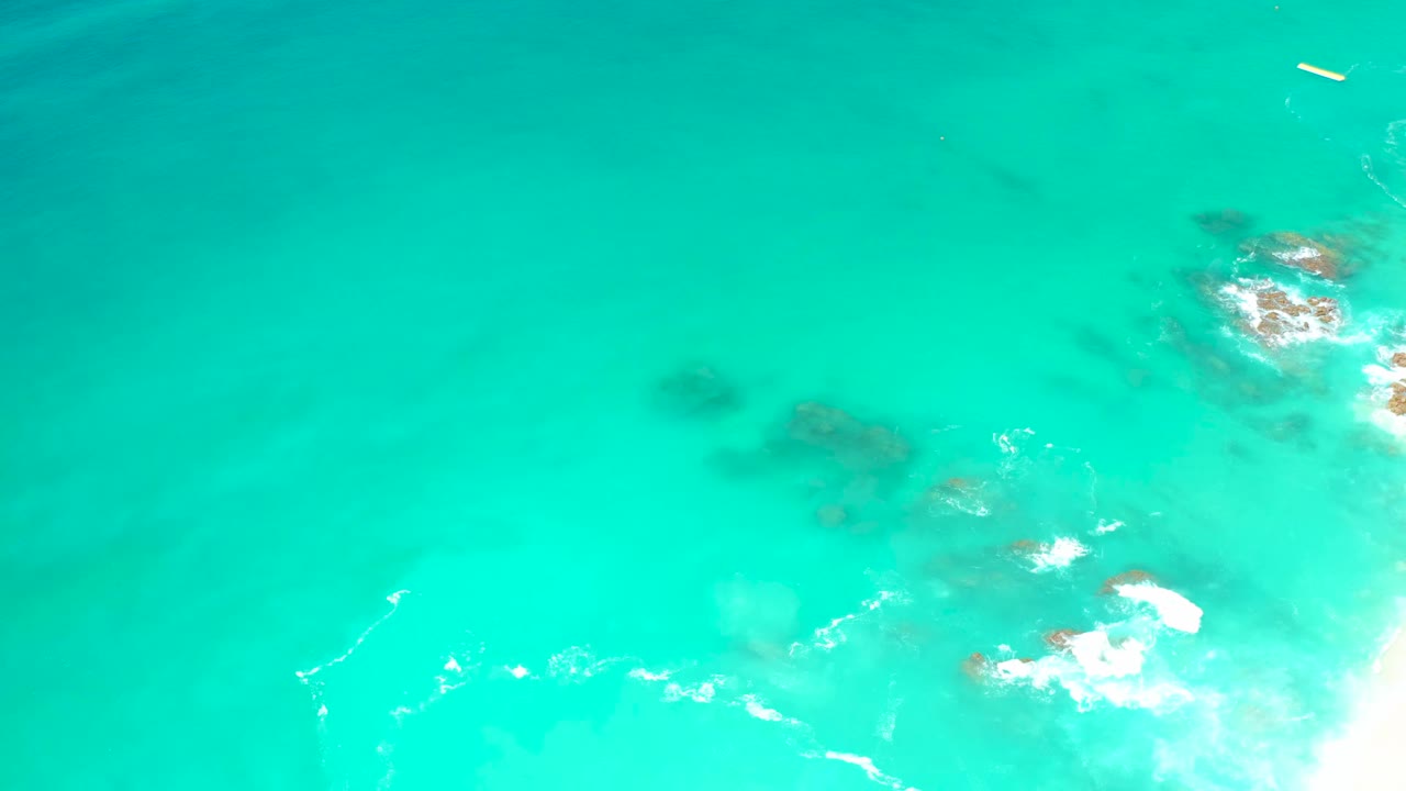 Aerial frame at Dreams Los Cabos showing clear turquoise water, darker submerged rock shapes, foam near the right edge, and no visible people.