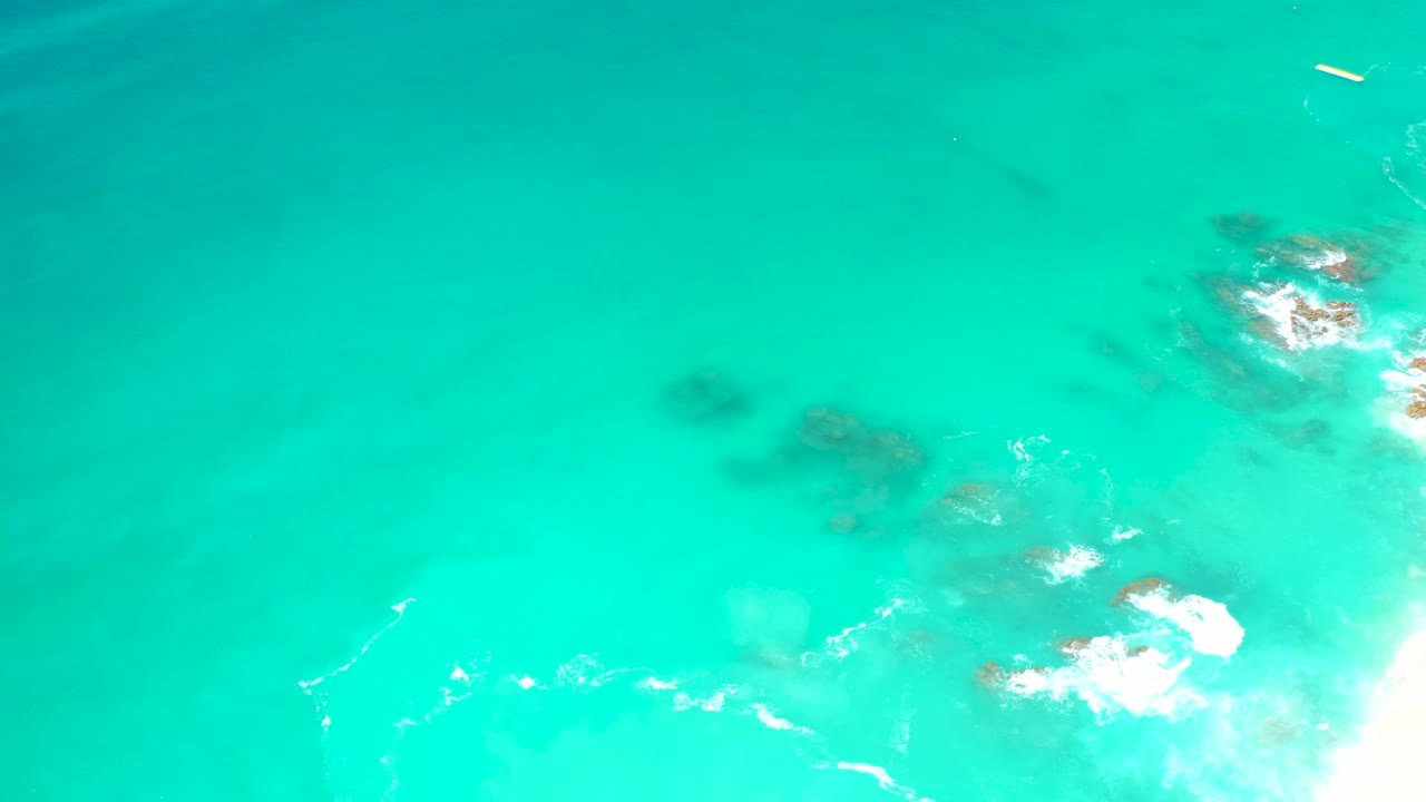 Overhead Los Cabos ocean detail with white foam spreading across partially visible rocks, open aqua water to the left, and no visible people.
