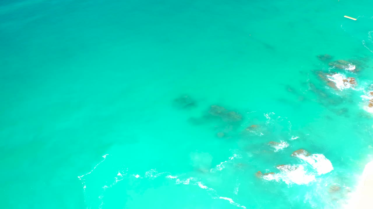 Top-down view at Dreams Los Cabos with bright turquoise water, rocky outcrops clustered to the right, fresh white foam, and no visible people.