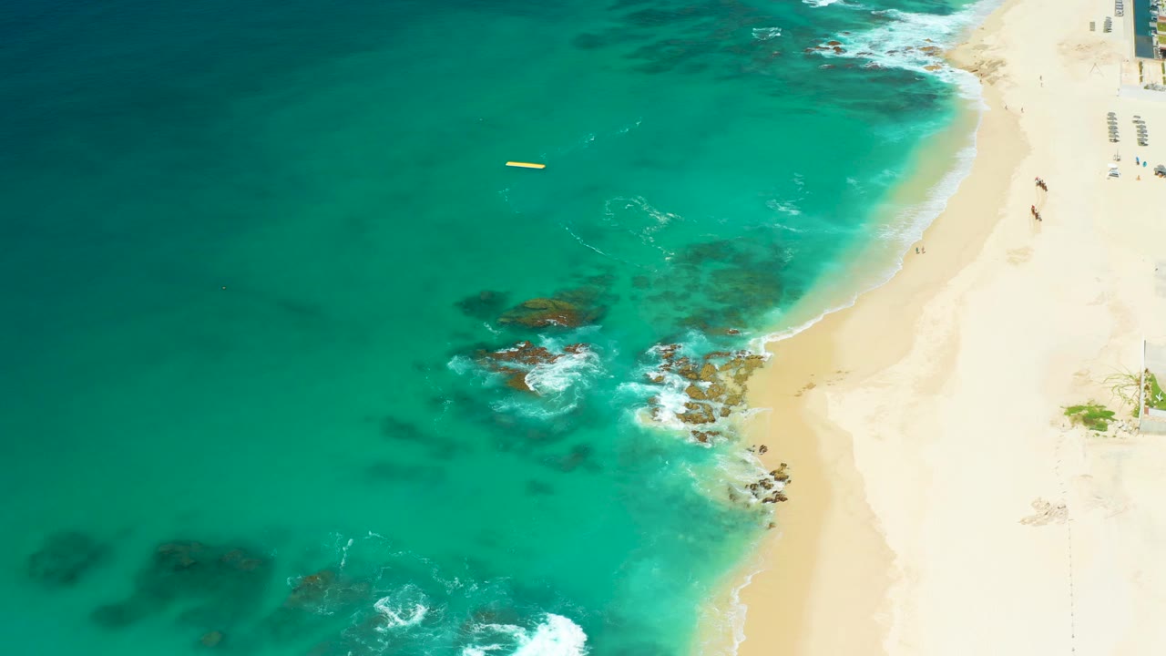 Overhead view at Dreams Los Cabos showing turquoise surf around dark rocks, pale sand along the right edge, a small yellow buoy offshore, and no visible people.