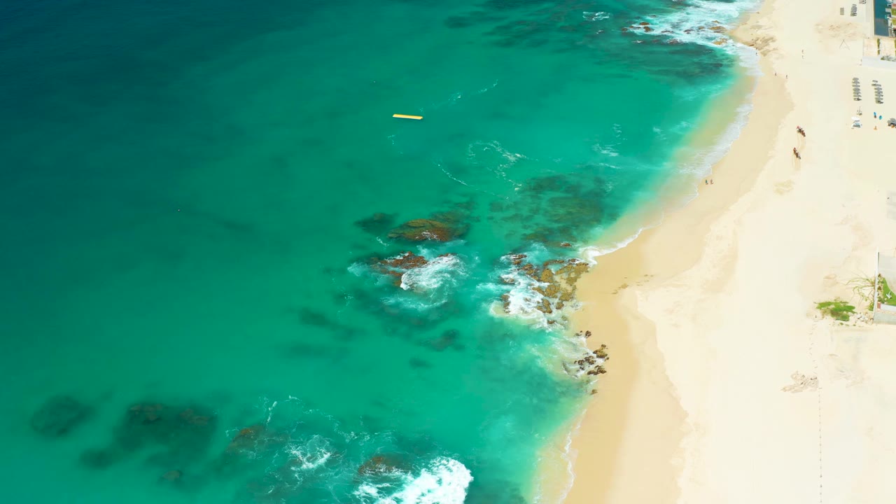 Top-down Dreams Los Cabos frame with a yellow buoy near the upper water, darker reef-like patches below the surface, rocks near the shore, bright sand, and no visible people.