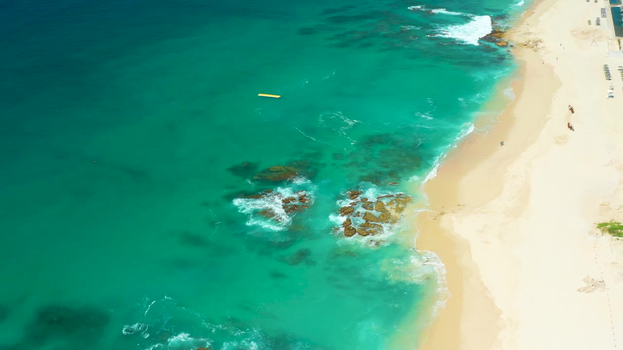 A high aerial frame at Dreams Los Cabos shows turquoise water on the left, pale beach on the right, rocky outcrops near the surf, a small yellow float offshore, and tiny people along the upper-right shore.