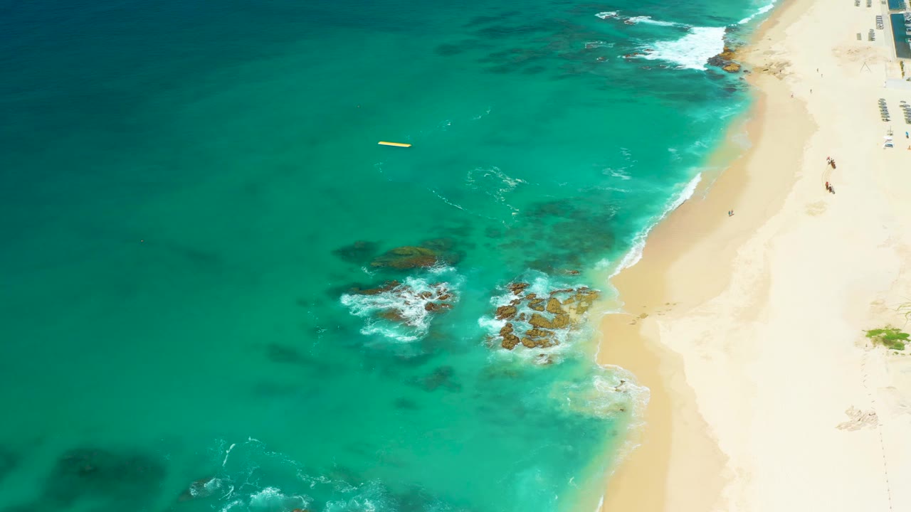 At 0:02, the overhead frame shows more of the pale sand on the right edge, blue-green water across the left, exposed rocks in the shallows, and tiny beachgoers near the upper-right shoreline.