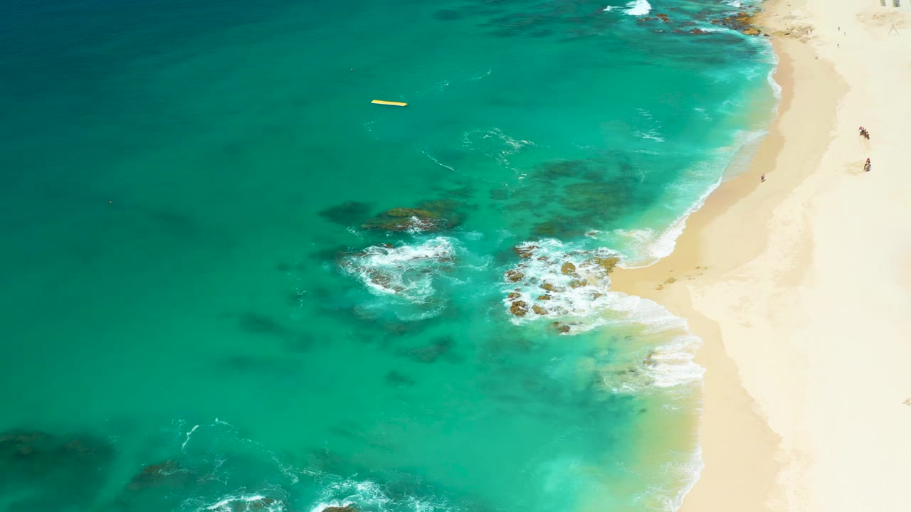 Turquoise surf meets rocks and sand with a yellow float offshore.