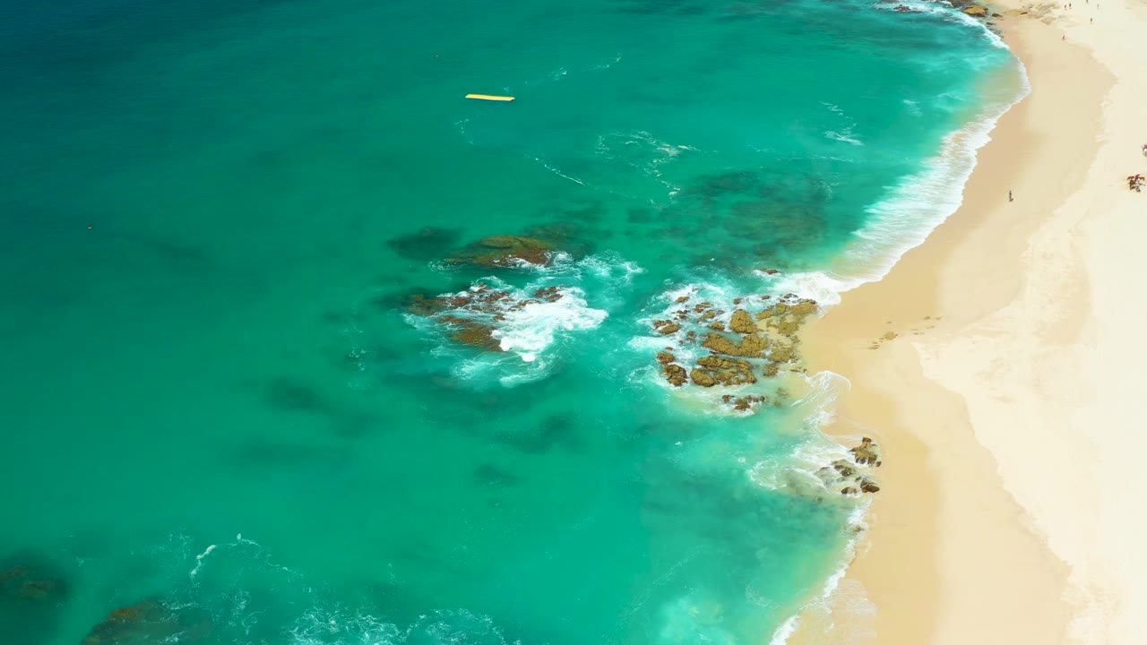 Aerial view at Dreams Los Cabos, Baja California Sur, Mexico, showing turquoise water, dark rocks, white surf foam, pale sand, a small yellow floating object offshore, and tiny people on the beach.