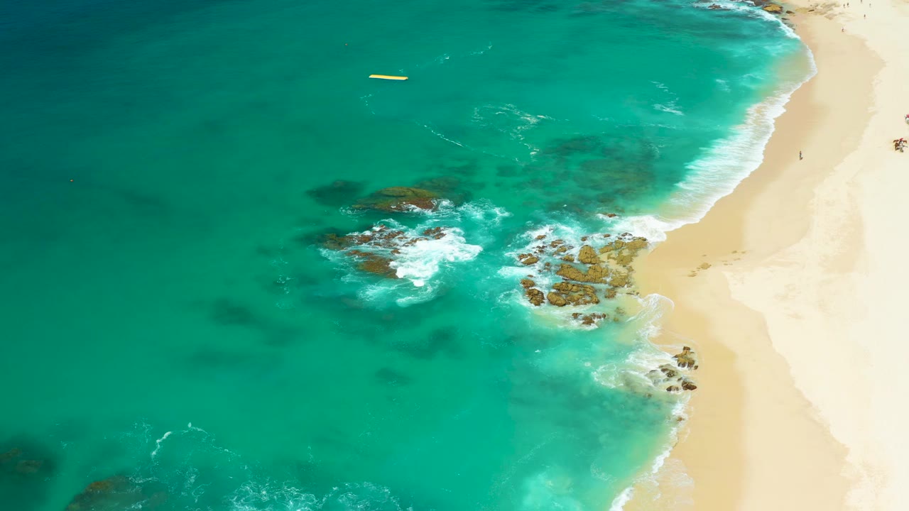 Overhead beach shoreline with turquoise water, rocks, foam, and a yellow object offshore.