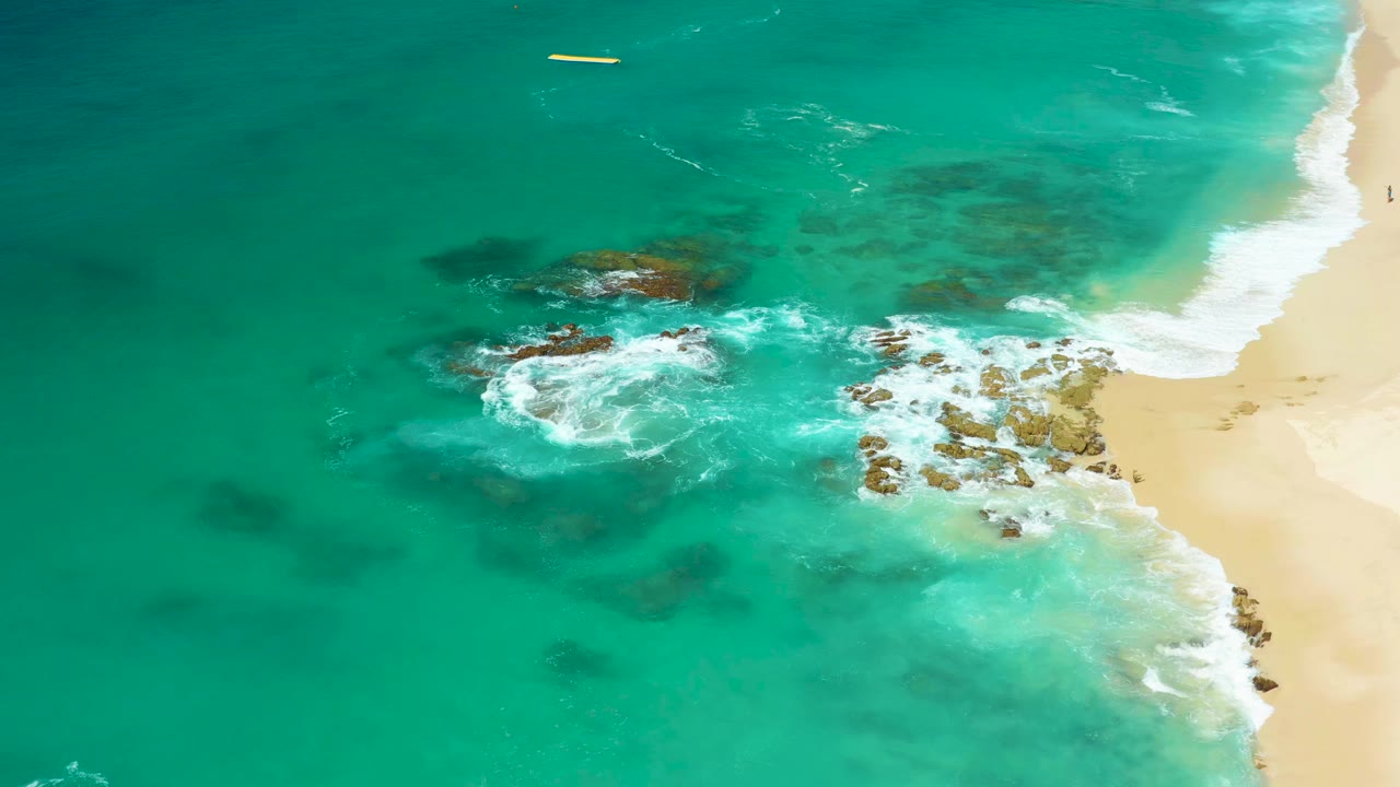 Overhead view at Dreams Los Cabos, Baja California Sur, Mexico, with turquoise water, white foam around rocky outcrops, pale sand on the right edge, a small yellow floating object offshore, and no visible people.