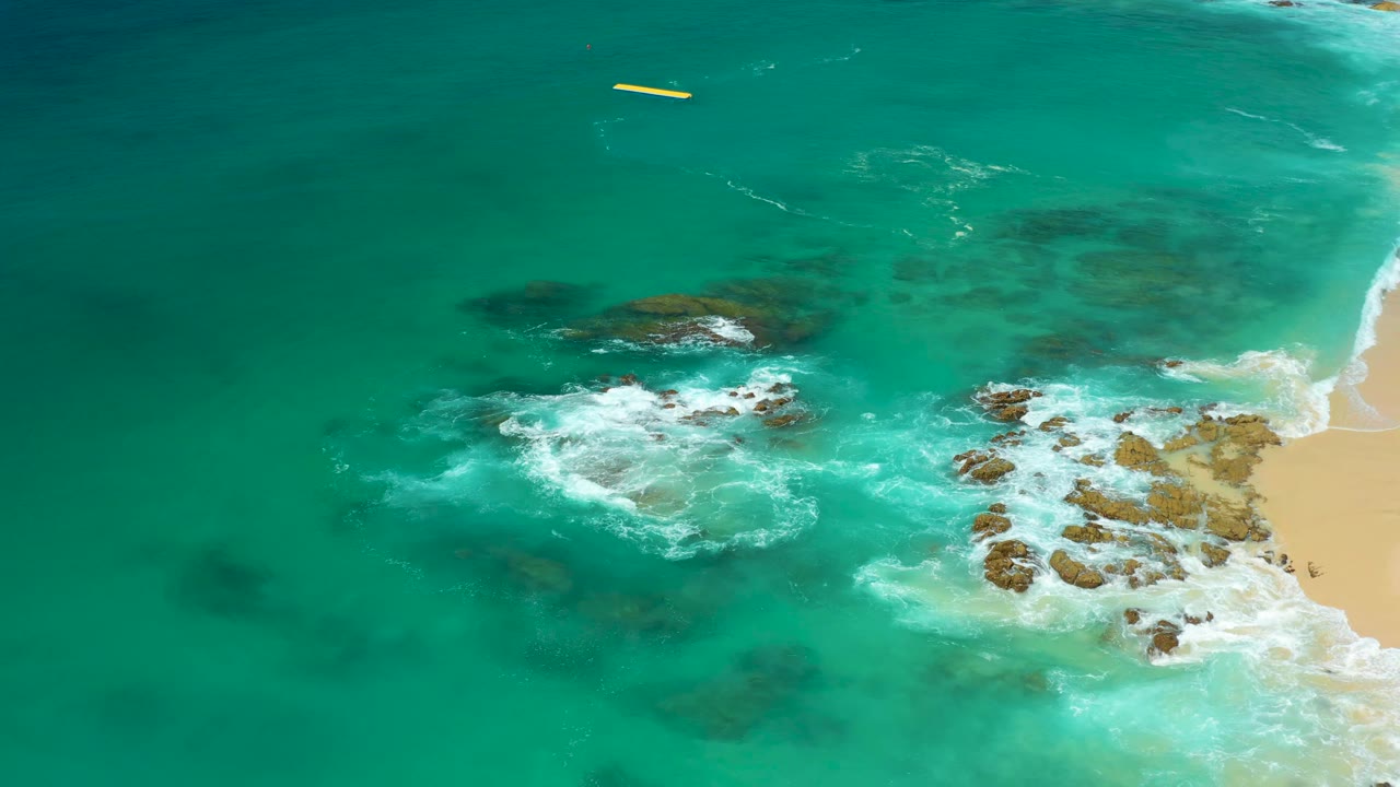Aerial frame from Dreams Los Cabos with foamy surf expanding around central rocks, darker underwater patches to the left, sand on the right, and no visible people.