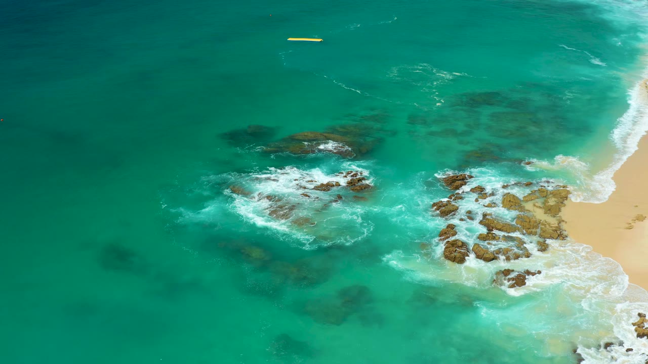 Dreams Los Cabos aerial frame showing a slightly lower shoreline position, with surf washing over rocks at center right, darker water patches at left, and no visible people.