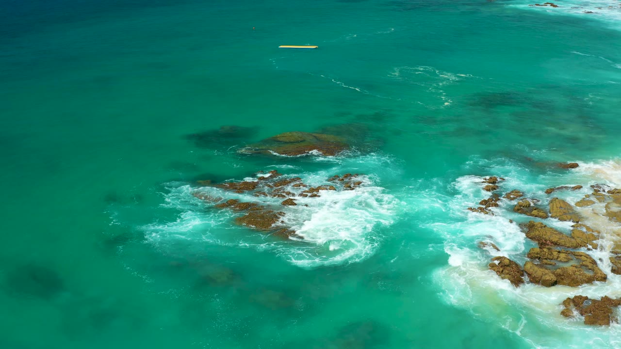 Aerial frame of turquoise water, white breaking waves, and rocky shore at Dreams Los Cabos, with sandy beach beginning at the right edge and no visible people.
