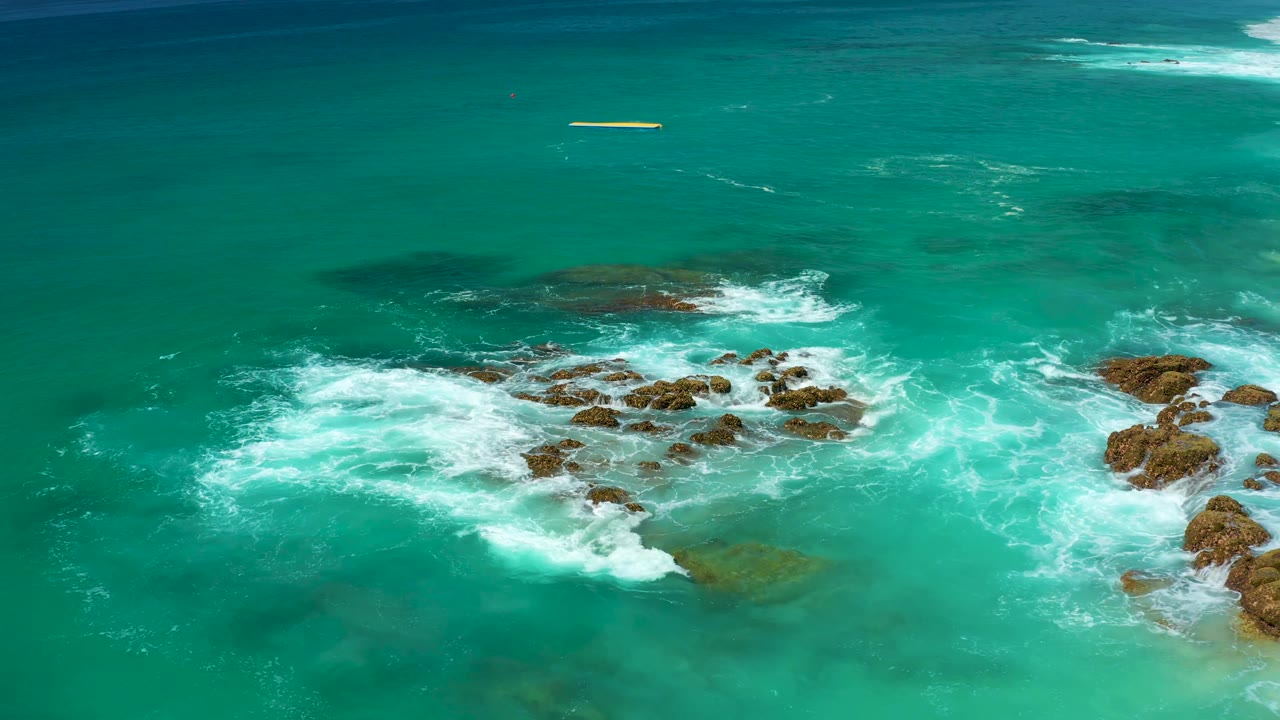 At 0:02, foamy water curls around scattered brown rocks while open turquoise water fills the upper frame, with no visible people.