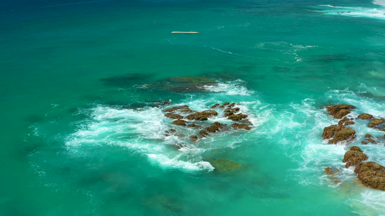 At 0:03, the aerial view places the strongest whitewater left of the main rocks, with clear green-blue water around them and no visible people.
