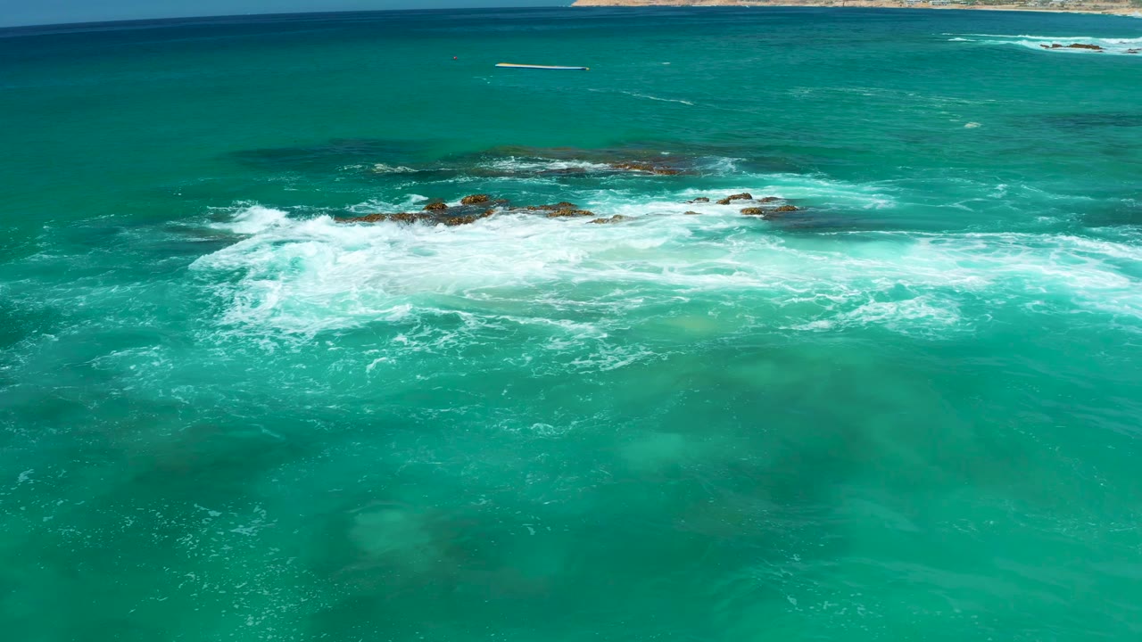 At Dreams Los Cabos in Baja California Sur, a broad band of white foam spreads across submerged rocks while deeper blue water sits beyond the break, with no visible people.