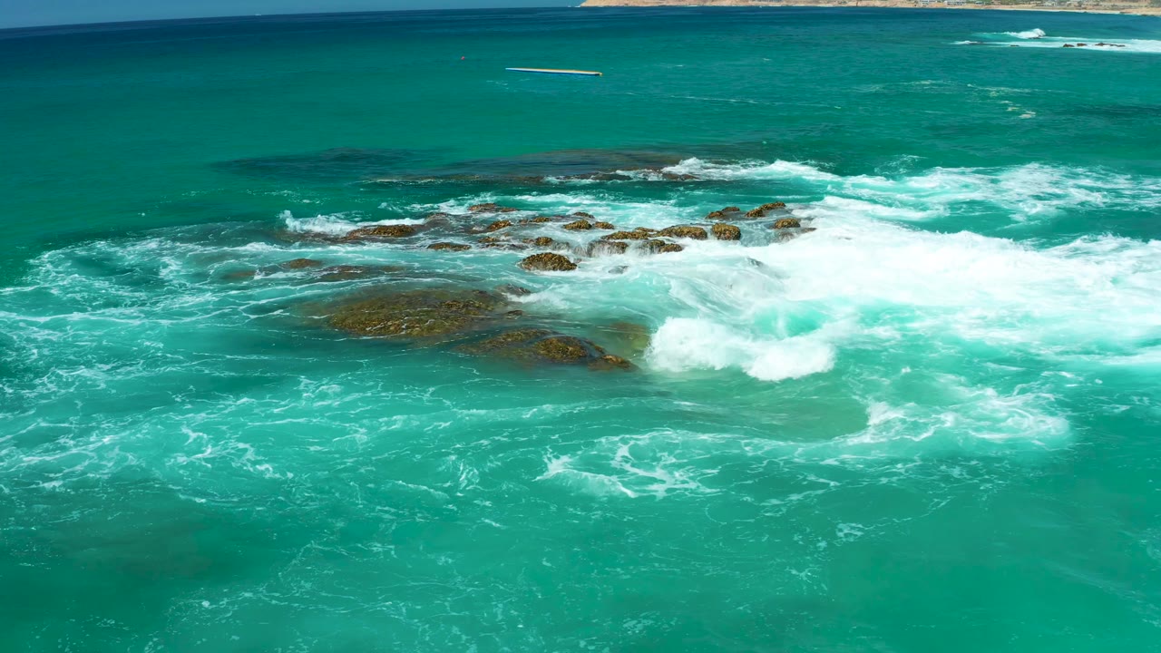 One second in, white surf expands across the central rocks at Dreams Los Cabos, with darker reef patches visible under the clear turquoise water and no visible people.