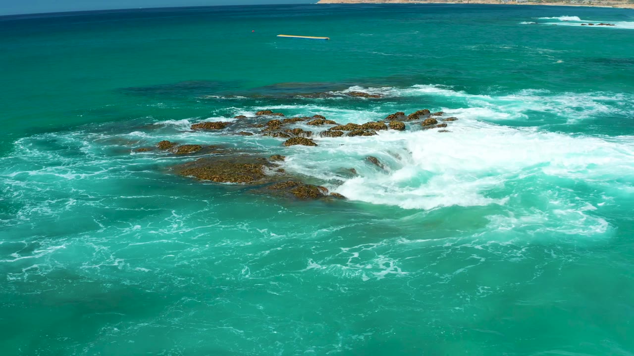 At 0:02, a broad white foam line rolls across low rocks while open turquoise water fills the foreground and a narrow coast remains along the top edge, with no visible people.