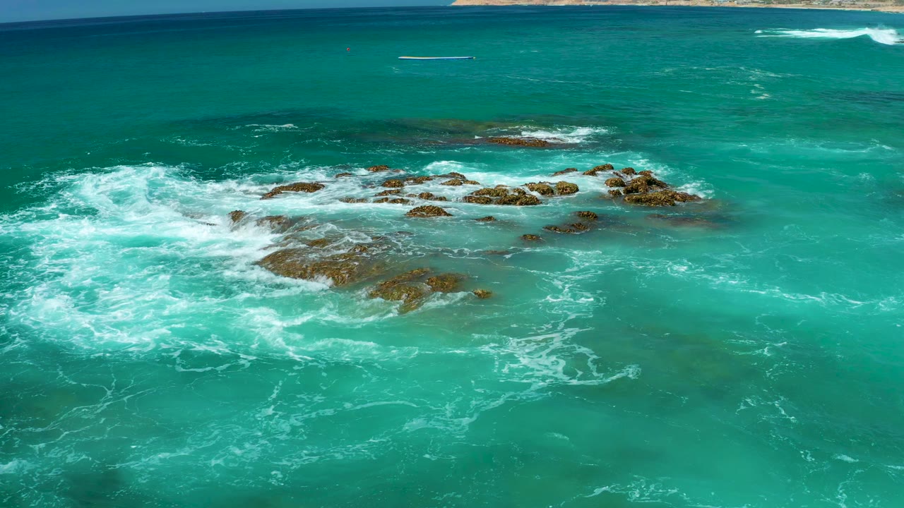 Aerial view at Dreams Los Cabos of turquoise water surrounding brown rocky outcrops as white surf spreads from the left, with no visible people.
