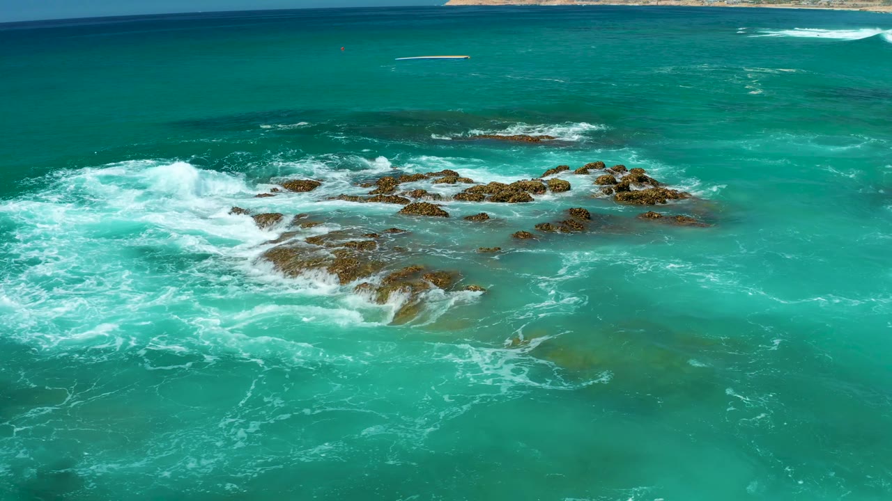 Aerial view at Dreams Los Cabos as a bright white wave crest breaks beside the midframe rocky formation while darker reef patches sit under the turquoise water, with no visible people.