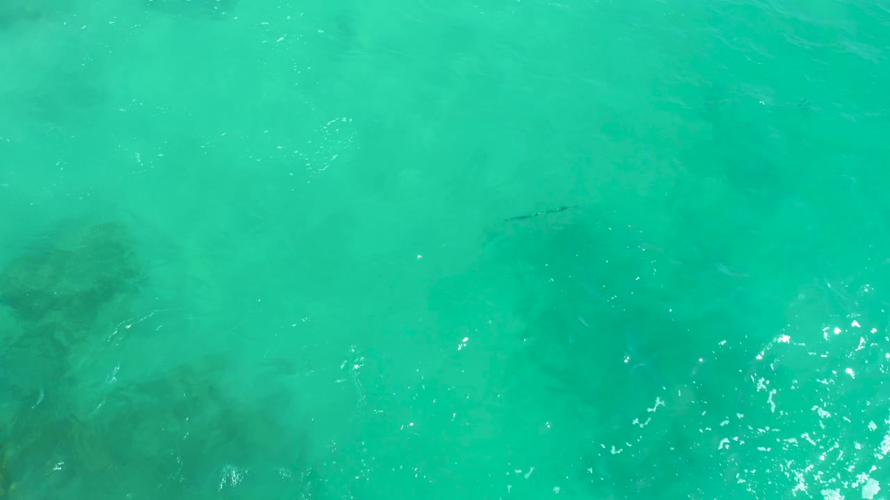 The overhead water view at Dreams Los Cabos becomes softer as ripples and surface haze partially obscure scattered submerged rocks, with no visible people.
