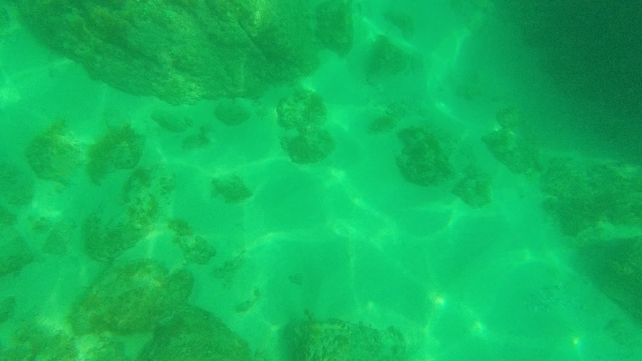 Underwater frame from Palmilla Los Cabos, Mexico showing darker shaded rocks, clear green water, pale sand between formations, and refracted sunlight, with no visible people.