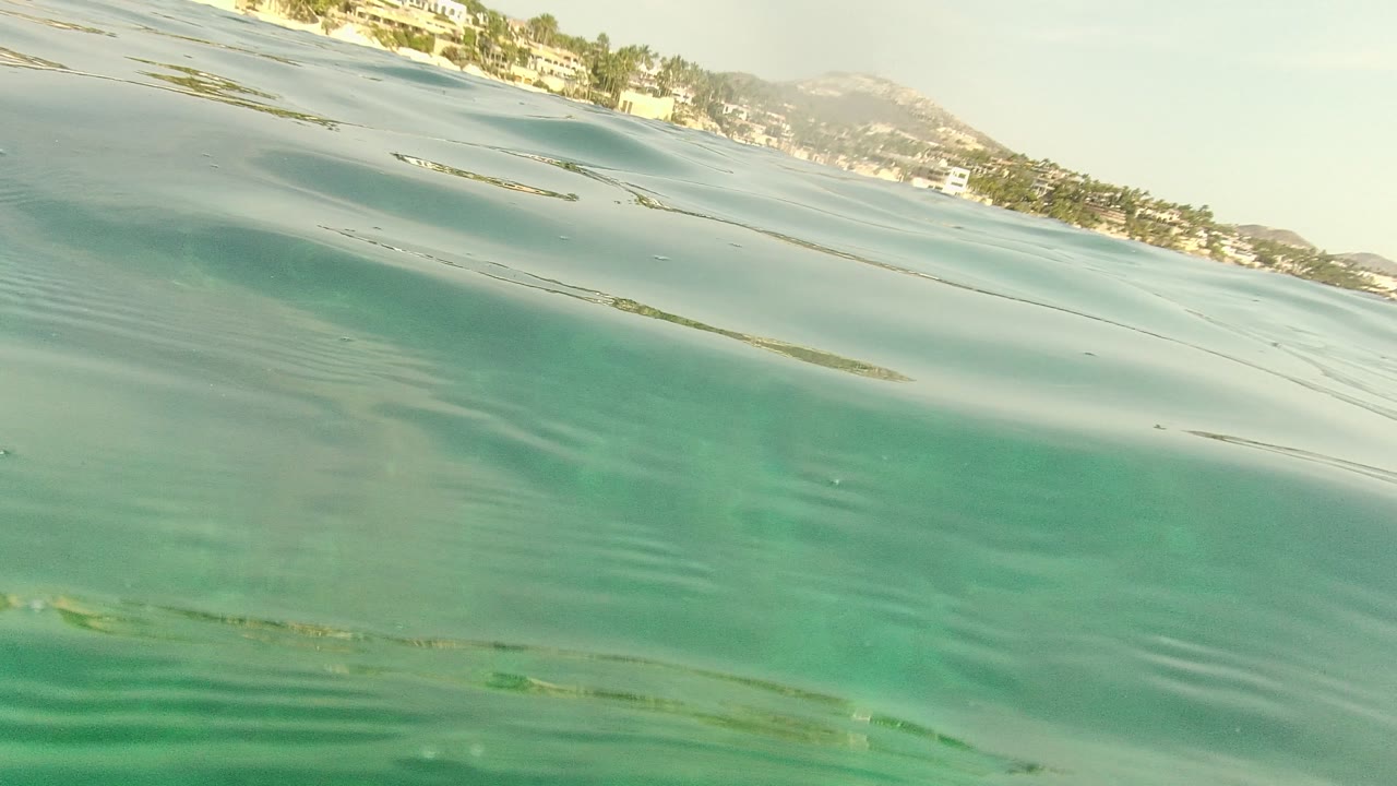 At Palmilla Los Cabos, the camera looks across rippled turquoise ocean toward a distant shore with buildings, trees, and low hills, with no visible people.