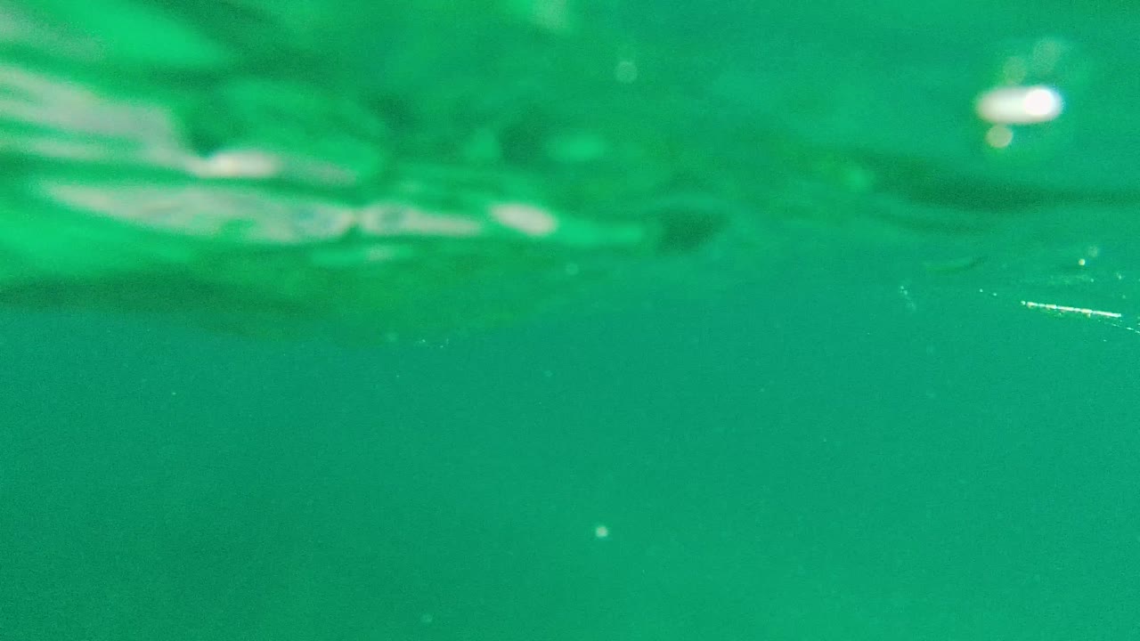 The camera has reached the ocean surface, showing teal water in front of the Palmilla Los Cabos shoreline, hillside buildings, distant hills, pale sky, and no visible people.