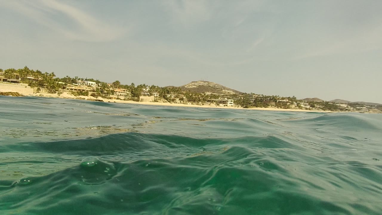 A blurred green swell fills the lower foreground while the Palmilla Los Cabos shoreline, buildings, hills, and pale sky remain visible above it, no visible people.