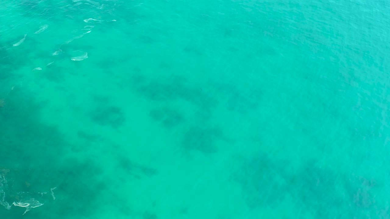 Top-down aerial frame from Shipwrecks Los Cabos with clear turquoise water, soft ripple texture, darker submerged patches toward the lower half, tiny boats near the upper left, and no visible people.