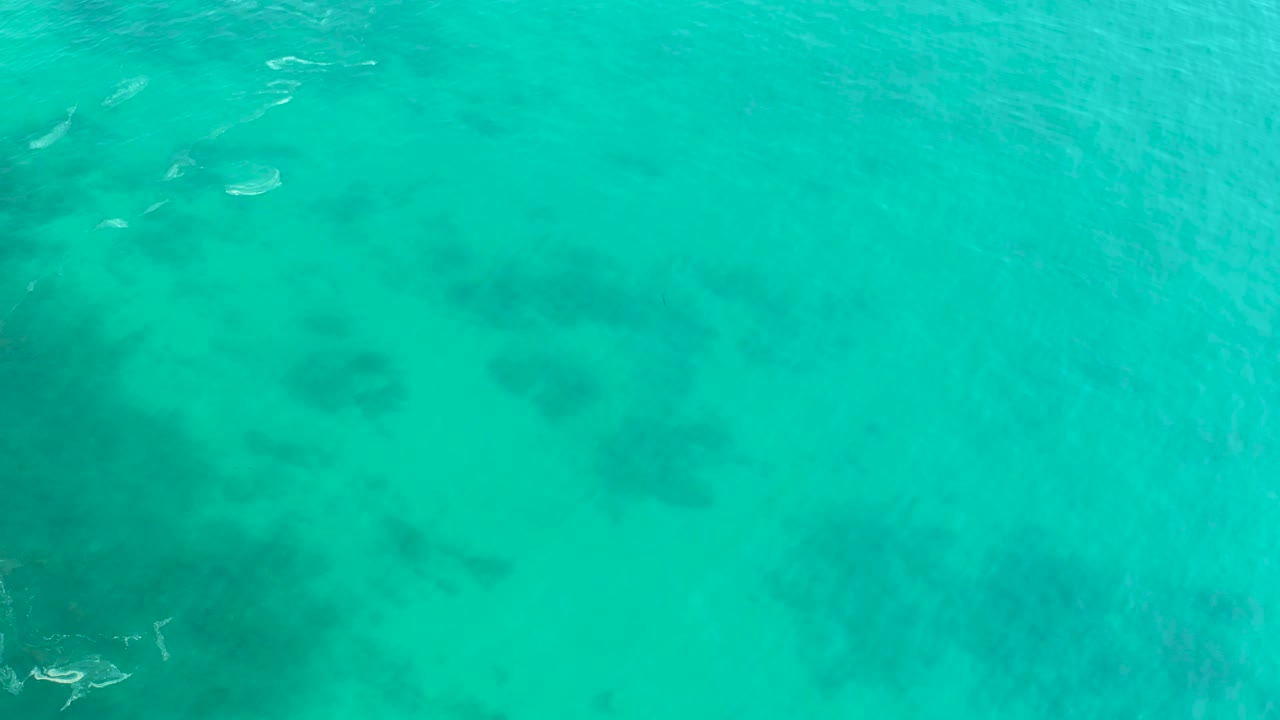 Aerial view over Shipwrecks Los Cabos with blue-green water, mottled darker formations below the surface, small boats near the top edge, and no visible people.