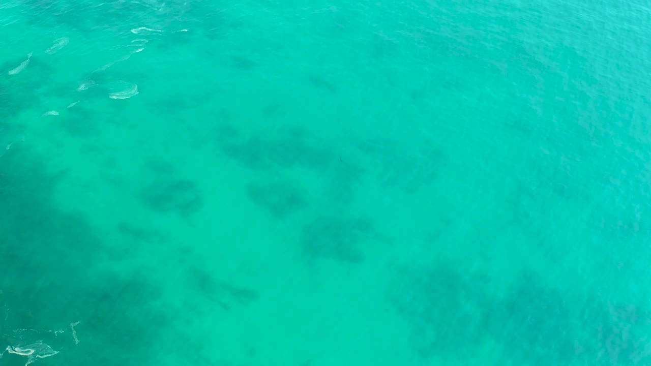 Overhead frame from Shipwrecks Los Cabos showing turquoise shallow water, dark underwater formations across the lower area, a small boat partly near the lower left edge, and no visible people.