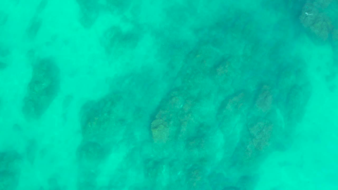 Aerial-looking top-down frame of turquoise sea with darker submerged rocks spread across the seabed at Shipwrecks Los Cabos, no visible people.