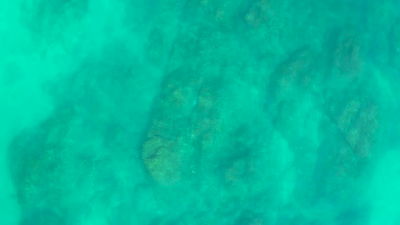 A darker cluster of submerged rocks is visible beneath turquoise water at Shipwrecks Los Cabos, with no visible people or vessels.